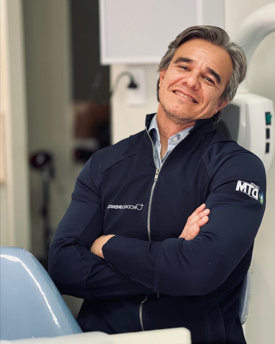 Dr. Rodrigo Wendel de braços cruzados, ao lado da cadeira do próprio consultório